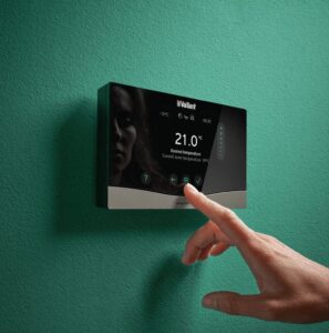 Primo piano del termostato intelligente Vaillant sensoCOMFORT, montato su una parete verde, con una mano che regola la temperatura impostata a 21°C